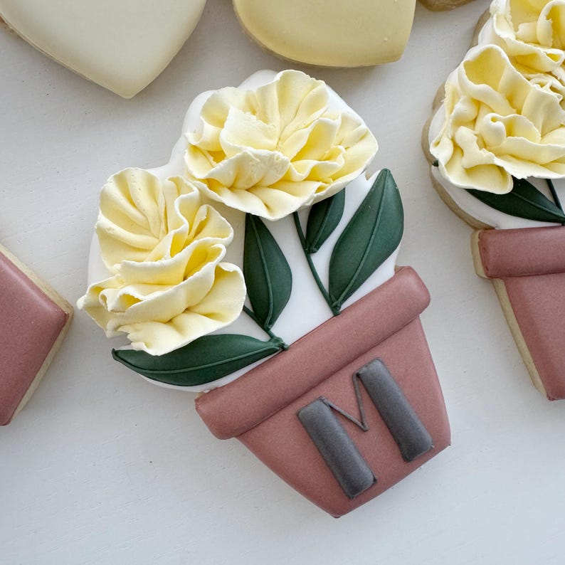 Peut inclure: Un biscuit d&eacute;cor&eacute; en forme de pot rose avec des fleurs jaunes. Le pot porte la lettre "M".