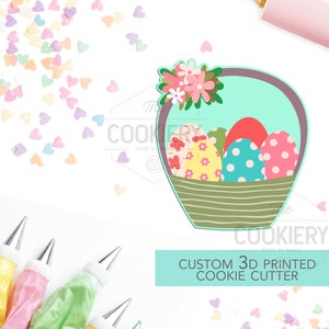 May include: A 3D printed cookie cutter shaped like a basket filled with Easter eggs. The basket is decorated with flowers and the eggs are decorated with polka dots and floral patterns. The cookie cutter is made by The Cookiery.