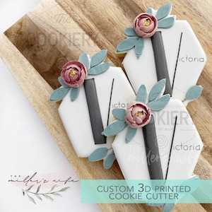 May include: Custom 3D printed cookie cutter in the shape of a hexagon with the letter "V" in the centre. The cutter is decorated with a pink flower and green leaves. The text "Victoria" is written on the cutter.