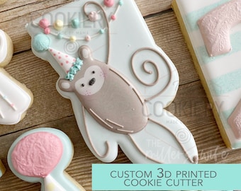 May include: A 3D printed cookie cutter shaped like a monkey wearing a party hat and holding a string of colourful balloons. The cutter is made of white plastic and has a light blue background. The text "CUSTOM 3D PRINTED COOKIE CUTTER" is printed on the bottom of the image.