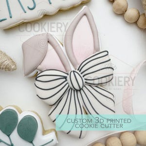 May include: A custom 3D printed cookie cutter shaped like bunny ears and a bow. The bow is white with black stripes. The bunny ears are pink with a light pink interior. The text "The Cookiery" and "Made by Mama" are visible.