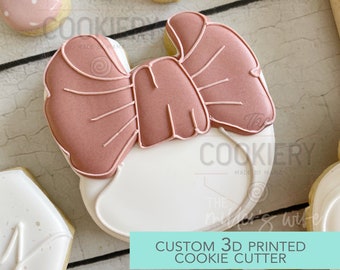May include: A 3D printed cookie cutter in the shape of a baby onesie with a pink bow on top. The cookie cutter is made of white plastic and has the text "The Cookiery Made by Mama" on the bottom.