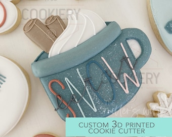 Puede incluir: Una galleta decorada con forma de taza de chocolate caliente con crema batida y palitos de canela. La taza es de color turquesa con la palabra "SNOW" en blanco, rosa y turquesa. Otras galletas en el fondo incluyen copos de nieve y un guante.