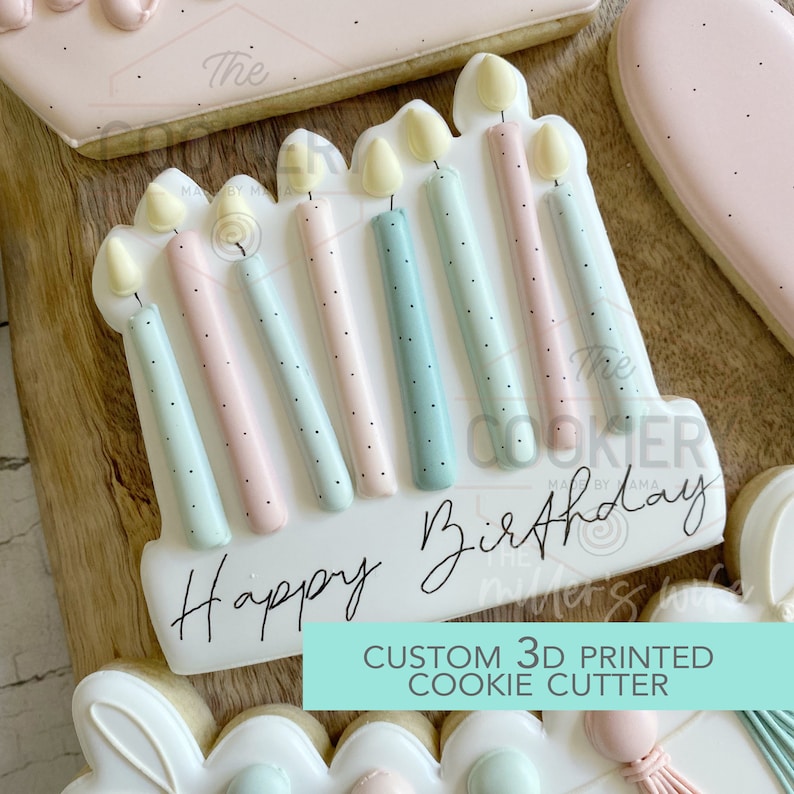 May include: A white cookie decorated with pastel blue, pink, and yellow icing. The cookie is shaped like a cake with candles on top. The text "Happy Birthday" is written on the cookie. The cookie is a custom 3D printed cookie cutter.