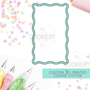 Op de afbeelding: Een lichtblauwe, golvende rechthoekige koekjessnijder. De afbeelding bevat een deegroller, spuitzakken en hartvormige hagelslag. De tekst luidt "The Cookiery" en "Custom 3D Printed Cookie Cutter". Bakbenodigdheden worden getoond.