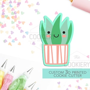 Könnte beinhalten: Ein individueller 3D-gedruckter Ausstecher in Form einer lächelnden Sukkulentenpflanze in einem gestreiften Topf. Der Ausstecher ist grün und rosa, mit dem Text "The Cookiery Made by Mama". Streusel und Spritzbeutel sind im Hintergrund.