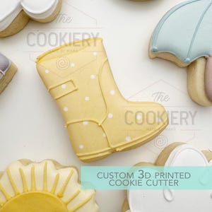 May include: A yellow cookie in the shape of a wellington boot, decorated with white polka dots. Other cookies, including an umbrella and a sun, are visible. The image includes the text "CUSTOM 3D PRINTED COOKIE CUTTER". The Cookiery logo is also present.