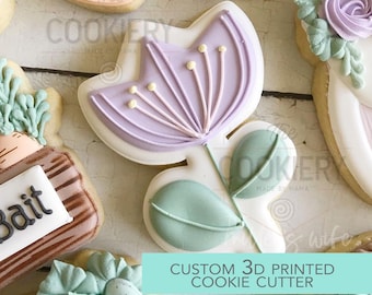 May include: A 3D printed cookie cutter in the shape of a purple flower with green leaves. The text "CUSTOM 3D PRINTED COOKIE CUTTER" is printed on a turquoise background.