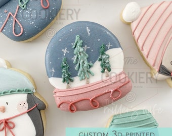 Pode incluir: Um cortador de biscoitos impresso em 3D com a forma de um globo de neve com uma base rosa e uma cúpula azul. O globo de neve tem três árvores verdes e neve branca dentro. O cortador de biscoitos é um design personalizado e é perfeito para fazer biscoitos de Natal.