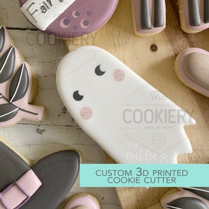 May include: A white ghost cookie cutter with black eyes and pink cheeks. The cookie cutter is on a wooden surface with other cookies in the background. The text "CUSTOM 3D PRINTED COOKIE CUTTER" is in the bottom right corner of the image.