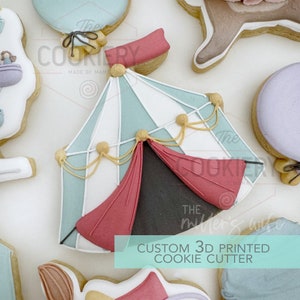 Op de afbeelding: Een 3D-geprinte koekjesvormer in de vorm van een circus tent met rode en blauwe strepen, gouden versiering en rode kleppen. De tekst "CUSTOM 3D PRINTED COOKIE CUTTER" is onder de tent gedrukt.