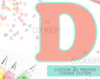 May include: A pink and green 3D printed cookie cutter in the shape of the letter D. The cookie cutter is on a white background with colorful heart sprinkles and piping bags.