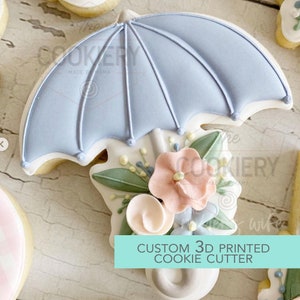 May include: A 3D printed cookie cutter in the shape of an umbrella with flowers. The umbrella is light blue with white trim and the flowers are pink, white, and green. The text "CUSTOM 3D PRINTED COOKIE CUTTER" is printed on a teal background.