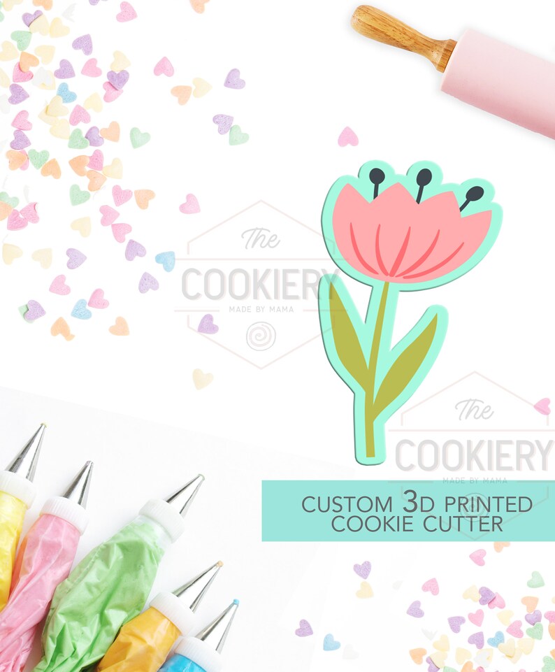 May include: A pink and green 3D printed cookie cutter shaped like a flower. The cutter is on a white surface with sprinkles and piping bags.