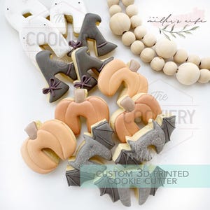 May include: A selection of Halloween-themed biscuits, featuring pumpkin, witch boot, and bat shapes. The biscuits are decorated with orange, grey, and brown icing. The image also includes text that reads "CUSTOM 3D PRINTED COOKIE CUTTER".
