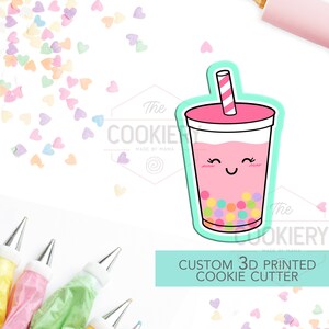 Op de afbeelding: Een op maat gemaakte 3D-geprinte koekjesvormer in de vorm van een roze bubble tea met een glimlachend gezicht en kleurrijke boba-parels. De vormer bevindt zich op een witte achtergrond met sprankels en spuitzakken.