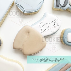 May include: A collection of custom 3D printed cookie cutters. One cookie cutter is shaped like a baby's bottom, with the text "Coming Oct '26". Other cookies are shaped like baby clothes. The cookies are beige, white, and light blue.