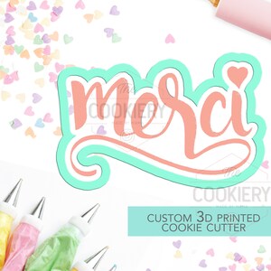 May include: A turquoise blue 3D printed cookie cutter in the shape of the word "Merci" with a small pink heart on the "i". The cookie cutter is on a white background with pink, yellow, and white heart sprinkles.