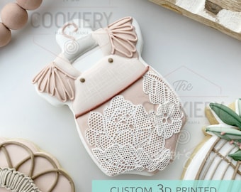May include: A 3D printed cookie cutter in the shape of a baby onesie with lace detailing. The onesie is pink and white with a brown button detail. The cookie cutter is on a white surface with a wooden background.