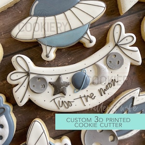 May include: A white cookie cutter shaped like a banner with the text "Two the Moon" and space-themed decorations, including a star, a crescent moon, and a planet. The cookie cutter is a custom 3D printed design.