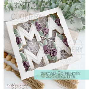 May include: A white box with a clear window showing three 3D printed cookie cutters in the shape of the letters "M-A-M" decorated with purple flowers and green leaves. The box is labeled "CUSTOM 3D PRINTED COOKIE CUTTER".