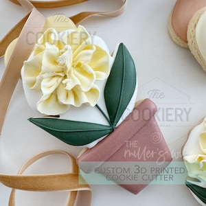 Peut inclure: Gros plan sur des biscuits décorés sur le thème floral. Un biscuit présente une grande fleur jaune froissée avec des feuilles vertes. Un autre biscuit est un carré rose avec le texte "CUSTOM 3D PRINTED COOKIE CUTTER". Rubans en arrière-plan.
