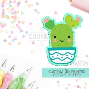 Könnte beinhalten: Ein grüner Kaktus-Ausstecher mit einem lächelnden Gesicht und zwei Armen. Der Kaktus befindet sich in einem blau-weißen Topf mit einem wellenförmigen Muster. Der Ausstecher ist 3D-gedruckt und ein individuelles Design. Das Bild zeigt außerdem einen Nudelholz, Spritzbeutel und Streusel.
