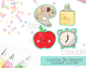 May include: A set of four 3D printed cookie cutters, featuring a paint palette, a glue bottle, a red apple with a smiling face, and a pink alarm clock. The cutters are made of white plastic and are designed for use with cookie dough. The image also includes a rolling pin, piping bags, and sprinkles.