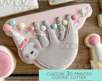 May include: A 3D printed cookie cutter shaped like a sloth hanging upside down with a party hat and a string of colourful beads. The text "CUSTOM 3D PRINTED COOKIE CUTTER" is printed on a turquoise background.