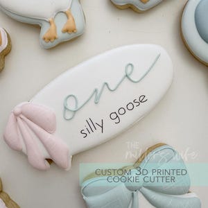 Puede incluir: Una galleta blanca con las palabras "one silly goose" en escritura cursiva. La galleta tiene una escritura azul claro y un lazo rosa en el lado izquierdo. Otras galletas son visibles en el fondo.