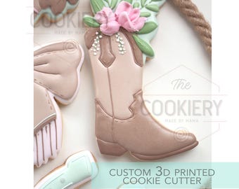 May include: A collection of decorated cookies, including a cowboy boot shape adorned with pink flowers and green leaves. The boot is light brown and beige. The text "CUSTOM 3D PRINTED COOKIE CUTTER" is visible.