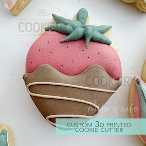 May include: A 3D printed cookie cutter shaped like a strawberry with chocolate dipped around the bottom. The strawberry has a green leafy top and is pink with white dots. The text "The Cookiery" is visible in the background.