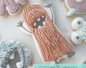 May include: A 3D printed cookie cutter shaped like a monster with long hair. The cookie cutter is made of white plastic and has a light brown color. The monster has a big mouth and two eyes. The text "CUSTOM 3D PRINTED COOKIE CUTTER" is written below the cookie cutter.