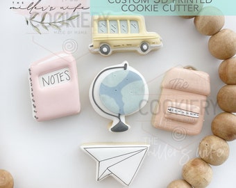 May include: Five 3D printed cookie cutters in the shape of a school bus, a globe, a notebook, a backpack, and a paper airplane. The cutters are on a white surface surrounded by wooden beads.