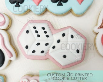 Puede incluir: Un cortador de galletas blanco y rosa con forma de dos dados conectados con puntos negros, que presenta un diseño central de dos dados de seis caras. El cortador tiene un borde rosa distintivo y secciones interiores blancas, con el texto The Cookiery.