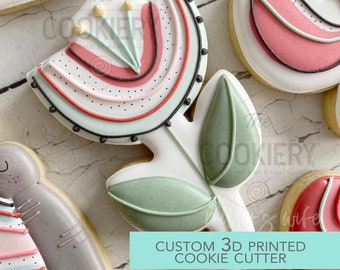 May include: A 3D printed cookie cutter in the shape of a stylized flower with a red, pink, and green design. The text "CUSTOM 3D PRINTED COOKIE CUTTER" is printed on a turquoise background.