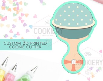 May include: A blue and white 3D printed cookie cutter shaped like a baby rattle. The rattle has a white ribbon and white polka dots on the blue part. The cookie cutter is on a white background with scattered pastel pink, blue, and yellow heart sprinkles.