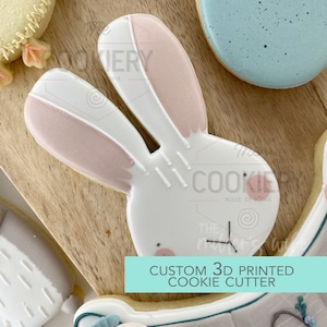 Può includere: Un tagliapasta a forma di coniglio bianco con orecchie rosa. Il tagliapasta è stampato in 3D e fa parte di un set di tagliapasta personalizzati. Il testo "CUSTOM 3D PRINTED COOKIE CUTTER" è nella parte inferiore dell'immagine.
