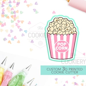 May include: A 3D printed cookie cutter in the shape of a popcorn container. The container is pink with white stripes and the word "POP CORN" in a white circle. The cutter is outlined in a light green color. The text "CUSTOM 3D PRINTED COOKIE CUTTER" is below the cutter.