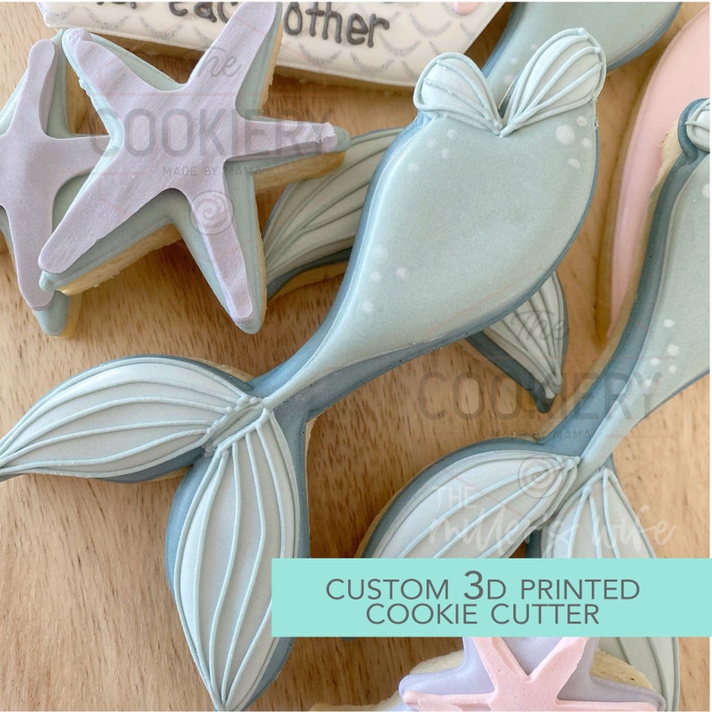 May include: A custom 3D printed cookie cutter in the shape of a mermaid tail. The cutter is light blue with white details and is shown on a wooden surface with other cookies.