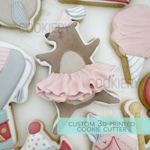 May include: A custom 3D printed cookie cutter shaped like a bear wearing a pink tutu and a party hat. The text "The Cookiery by Mama" is visible in the background.