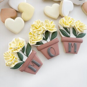 Peut inclure: Trois biscuits en sucre d&eacute;cor&eacute;s en forme de pots de fleurs avec des fleurs jaunes et des feuilles vertes. Les pots sont bruns et portent les lettres "M O M".