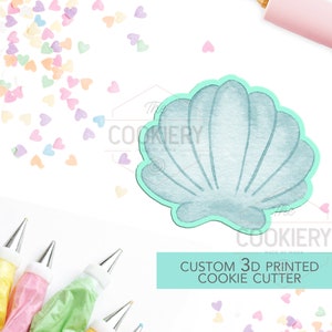 May include: A light blue 3D printed seashell cookie cutter with a textured surface. The cutter is on a white background with colourful heart sprinkles and piping bags.
