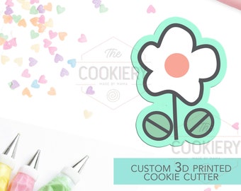 May include: A 3D printed cookie cutter in the shape of a flower with a pink center and two green circles at the bottom. The cutter is on a white background with colorful sprinkles.