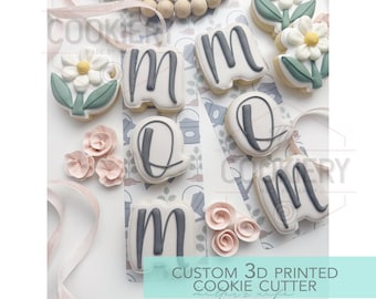 May include: An overhead shot of decorated cookies spelling "MOM" and flower-shaped cookies. The cookies are white with gray lettering and accents. The image also includes pink paper flowers, wooden beads, and the text "CUSTOM 3D PRINTED COOKIE CUTTER".