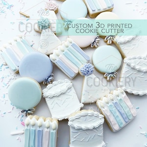 Puede incluir: Una variedad de galletas personalizadas impresas en 3D con formas de globos, velas y pasteles. Decoradas con glaseado pastel azul, blanco y rosa. El texto "CUSTOM 3D PRINTED COOKIE CUTTER" es visible. Dispuestas sobre una superficie blanca.