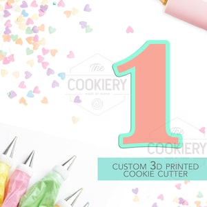 May include: A pink and green 3D printed cookie cutter in the shape of the number one. The cutter is on a white background with sprinkles and piping bags.
