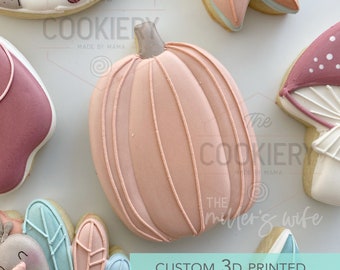 May include: A 3D printed cookie cutter shaped like a pumpkin. The cutter is pink with white lines and a stem. The text "CUSTOM 3D PRINTED COOKIE CUTTER" is at the bottom of the image.