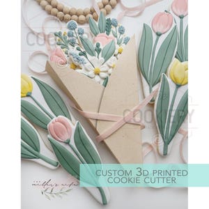 Op de afbeelding: Een verzameling op maat gemaakte 3D-geprinte koekjessnijders, waaronder tulp- en bloemontwerpen. De koekjes zijn versierd met pastelroze, gele en blauwe glazuur, en zijn gerangschikt met een lichtbruine papieren houder en roze lint.
