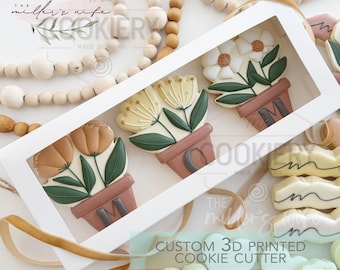 May include: Three 3D printed cookie cutters in the shape of potted flowers. The cutters are made of white plastic and are displayed in a white box with a clear window. The box has the text "CUSTOM 3D PRINTED COOKIE CUTTER" printed on the front.